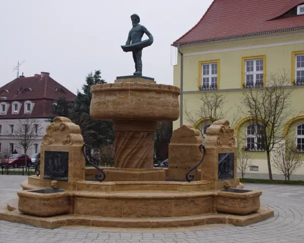 Kamienna fontanna z rzeźbą rybaka na szczycie, ozdobiona płaskorzeźbami statków.