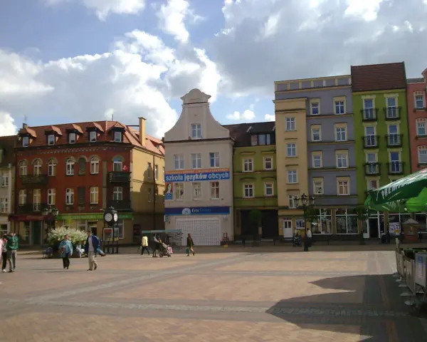 Rynek w Chojnicach z kolorowymi kamienicami, w tym ratusz. Ludzie spacerują po brukowanym placu.