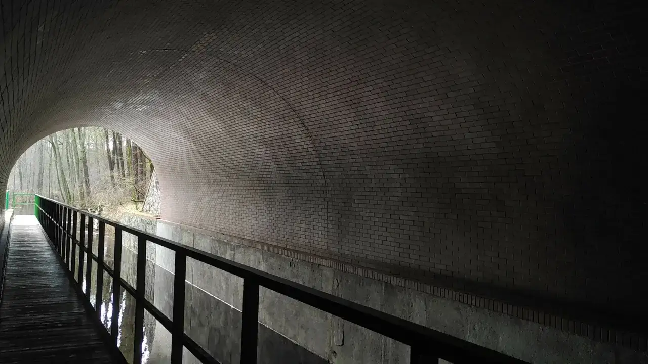 Wnętrze ceglanego tunelu, przez który biegnie ścieżka z balustradą. W oddali widać wyjście na las. To akwedukt w Fojutowie.
