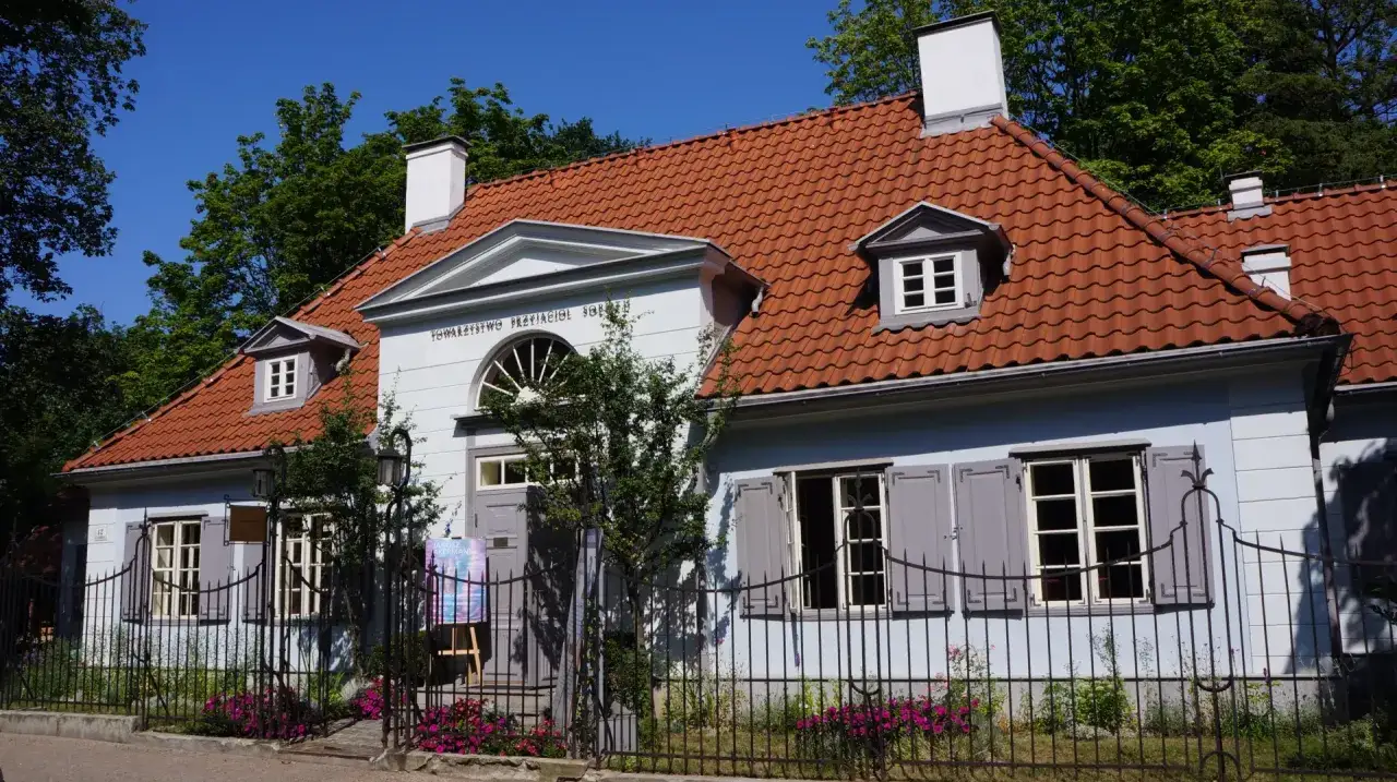 Jasnoniebieski dworek Sierakowskich z czerwoną dachówką, otoczony zielenią i ozdobnym płotem.