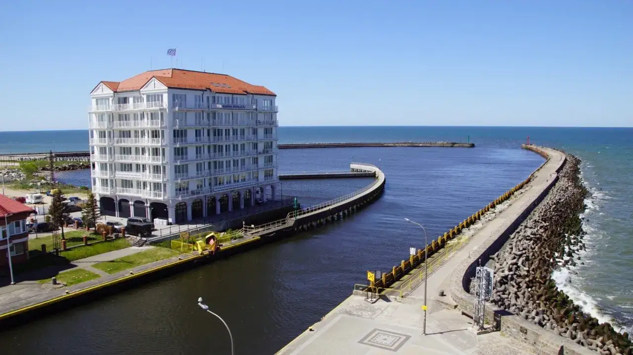 Nowoczesny budynek Marina Royale przy kanale portowym w Darłówku Zachodnim, z falochronami prowadzącymi do morza.