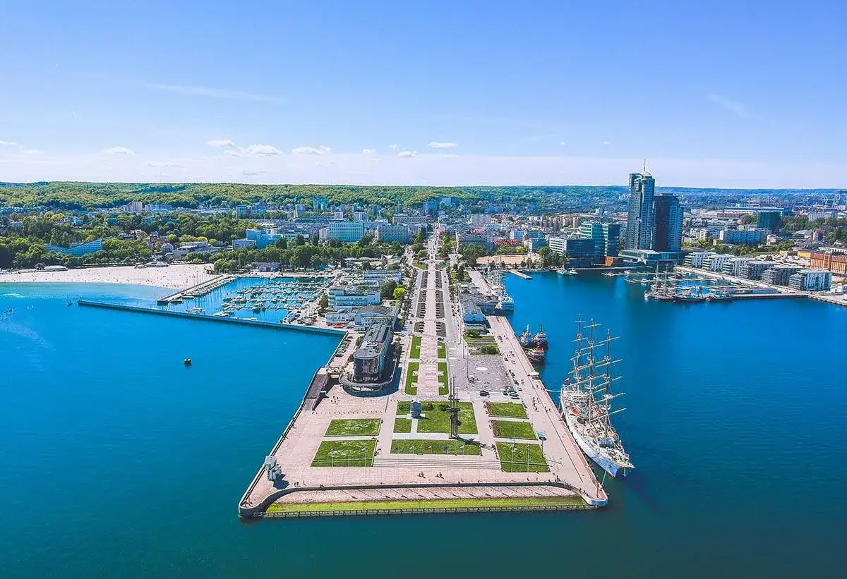 Widok na port i nowoczesne budynki Gdyni. Na pierwszym planie biały żaglowiec, a w tle plaża i zielone wzgórza. To piękna Gdynia, stare miasto i jego okolice.