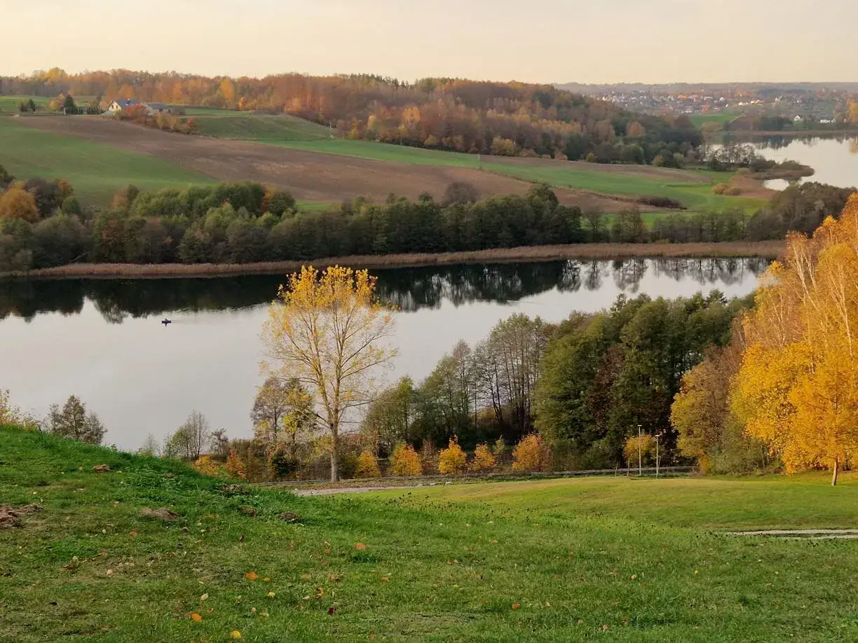 Spokojna jesień na Kaszubach. Jezioro odbija złote liście drzew, a w oddali widać pola i domy.