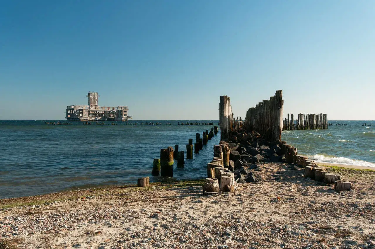 Ruiny betonowej budowli na morzu, przypominającej okręt, w oddali. Na pierwszym planie zniszczone drewniane pale i kamienie na plaży. Babie doły Gdynia.