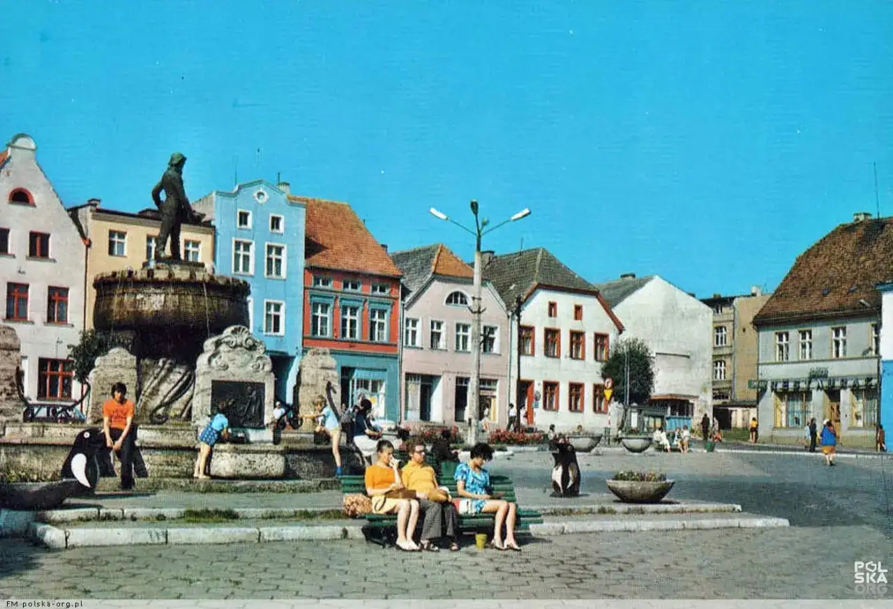 Pomnik rybaka góruje nad placem w otoczeniu kolorowych kamienic. Dzieci bawią się przy fontannie, a ludzie odpoczywają na ławkach.