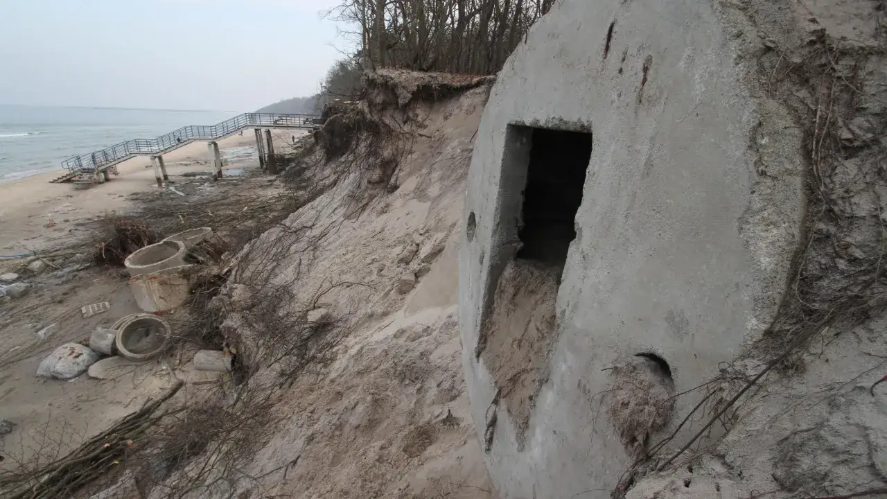 Zniszczone bunkry nad morzem, z widoczną drabiną prowadzącą na plażę i drzewami na skarpie.