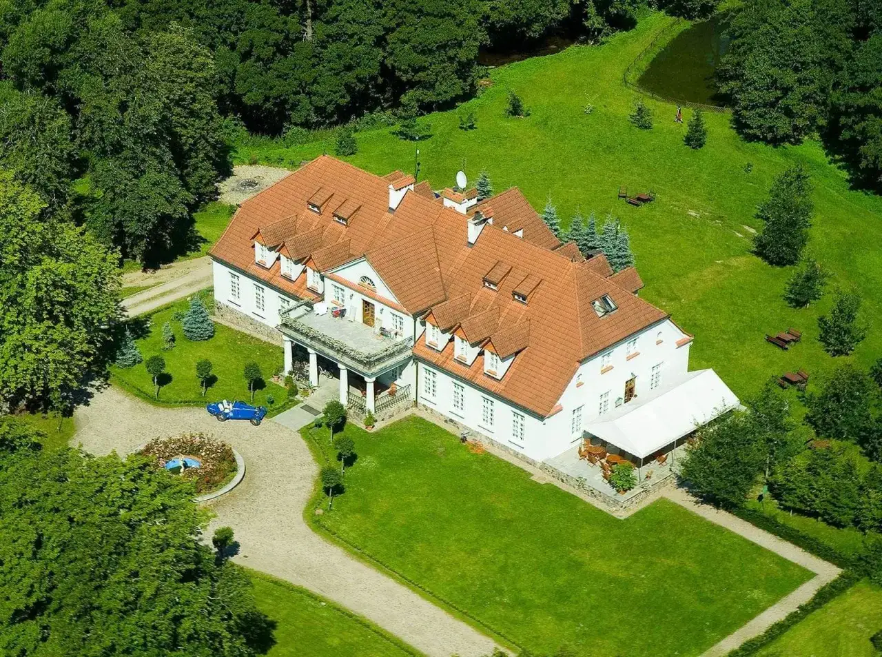 Dwór w Bychowie otoczony zielenią, z niebieskim kabrioletem przed wejściem.