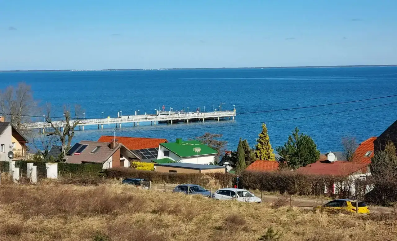 Widok na spokojne morze i molo w oddali. Na pierwszym planie domki z czerwonymi dachami i żółty samochód. Mechelinki plaża w słoneczny dzień.