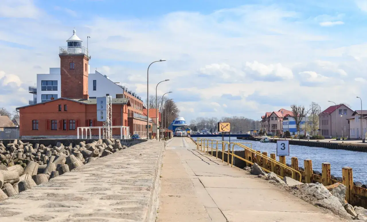 Latarnia morska i nabrzeże w Darłówku, z widokiem na kanał portowy i budynki.