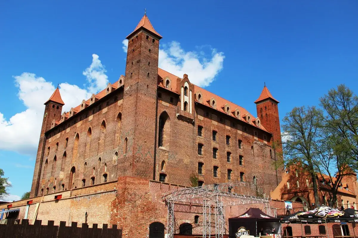 Gotycki zamek z czerwonej cegły, z wieżami i dachami, pod błękitnym niebem. W dole scena i namioty, zapowiadające atrakcje.