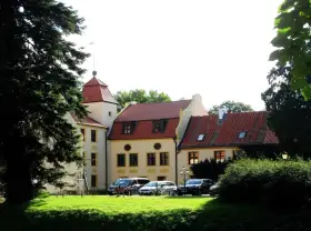 Zamek w Krokowej z czerwoną wieżą i dachami, otoczony zielenią. Przed budynkiem zaparkowane samochody.