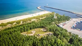 Widok z lotu ptaka na wybrzeże z piaszczystą plażą, lasem i portem. Wśród drzew widoczne są historyczne bunkry nad morzem.