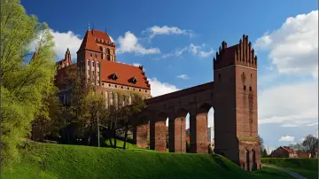 Majestatyczny zamek w Kwidzynie, z czerwonej cegły, z charakterystycznym gdaniskiem i długim, arkadowym murem obronnym, wznosi się na zielonym wzgórzu pod błękitnym niebem.