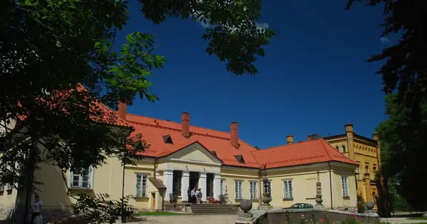Pałac w Waplewie Wielkim, żółty, z czerwonym dachem, otoczony zielenią, pod błękitnym niebem.