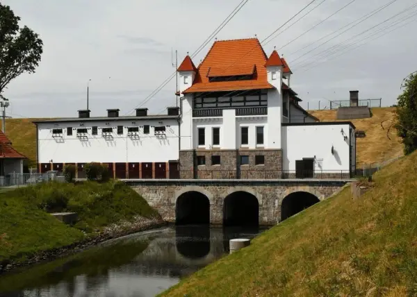 Elektrownia wodna Straszyn z charakterystycznym czerwonym dachem i kamiennymi arkadami nad kanałem.