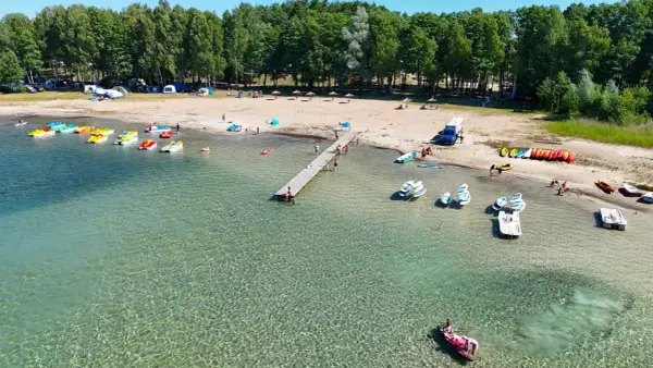 Letnia przygoda na wodzie, kolorowe rowerki wodne i kajaki czekają przy brzegu. Widok na plażę z namiotami i lasem, idealne miejsce na wypoczynek nad jeziorem Binduga 69.
