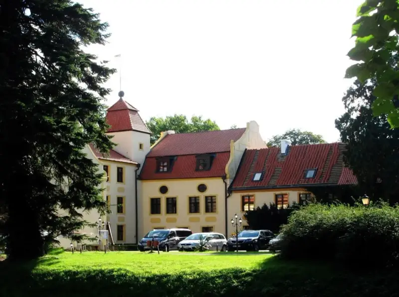 Zamek w Krokowej z czerwoną wieżą i dachami, otoczony zielenią. Przed budynkiem zaparkowane samochody.